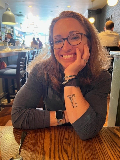 Photo of Jen smiling warmly at a restaurant table, resting her chin on her hand in a relaxed, friendly pose.