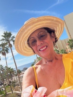 Photo of Yoli smiling outdoors in a sun hat and yellow top, with palm trees and a bright blue sky in the background.